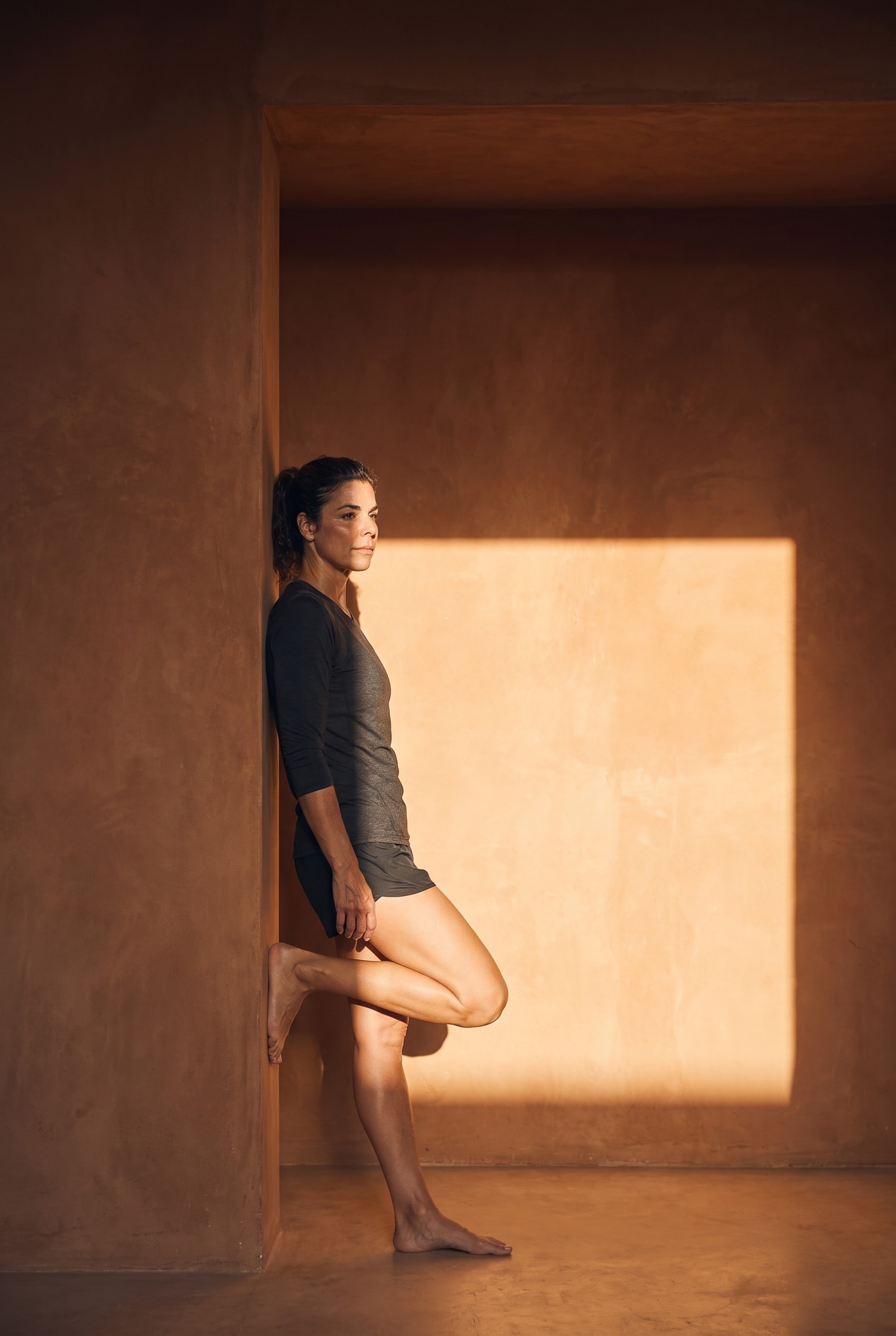 An athlete resting against a warm plaster wall with one foot raised, in hard sunlight.