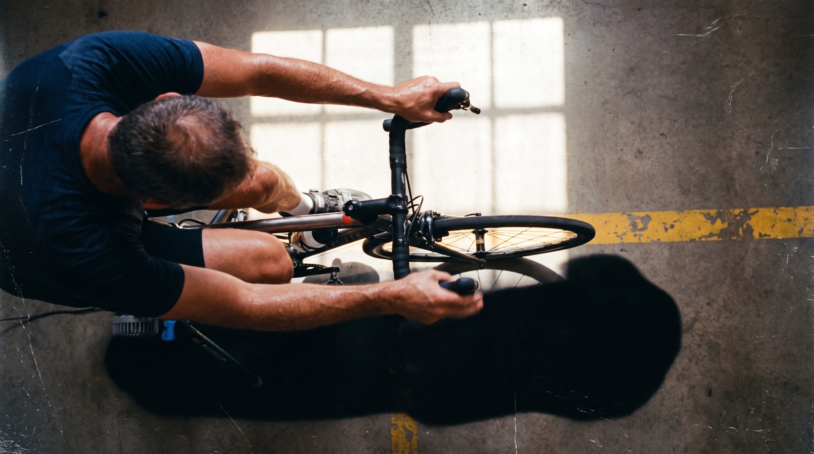 An overhead frame of a cyclist on an indoor trainer, head down over the bars in hard window light.